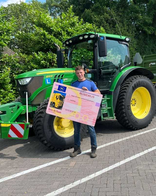Geslaagd voor het T-rijbewijs: cursist met rijbewijsbord naast een groene lestrekker met aanhangwagen in de zon