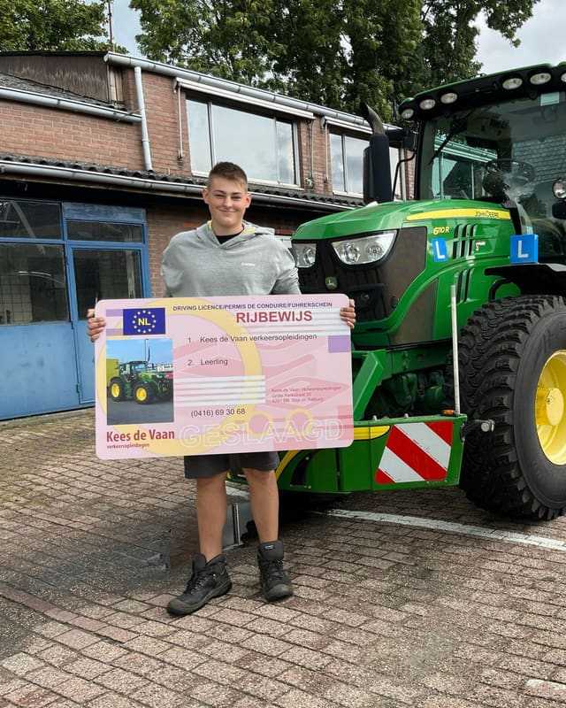 Geslaagd voor het trekkerrijbewijs: cursist in witte trui met groot rijbewijsbord voor het lesgebouw