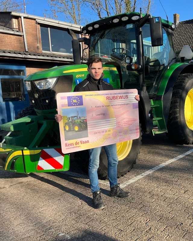 Geslaagd voor het trekkerrijbewijs: cursist met groot rijbewijsbord voor een groene lestrekker bij het lesgebouw