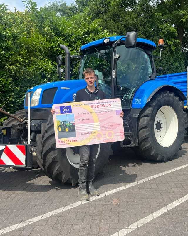 Geslaagd voor het trekkerrijbewijs: cursist met rijbewijsbord voor een blauwe lestrekker met blauwe aanhanger