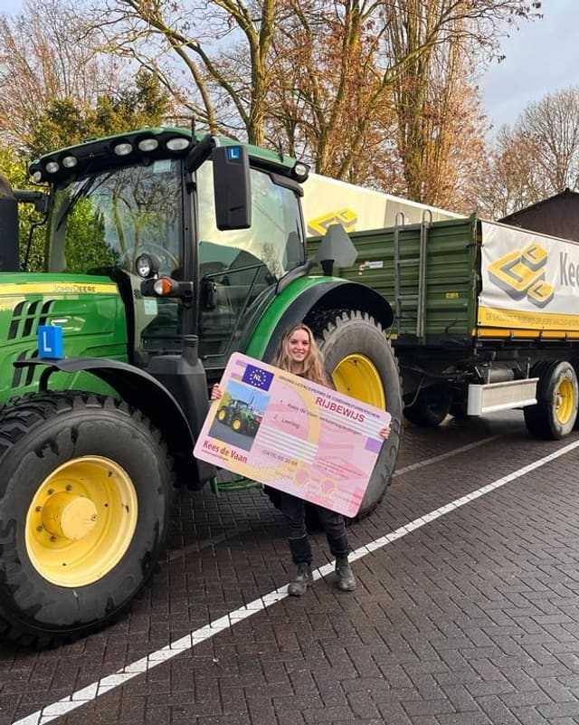 Geslaagde cursist met rijbewijsbord voor een groene lestrekker met aanhangwagen van Kees de Vaan