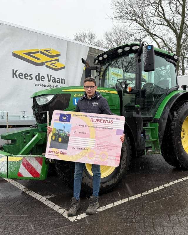 Geslaagde cursist met rijbewijsbord naast een groene lestrekker, met een lesvrachtwagen op de achtergrond