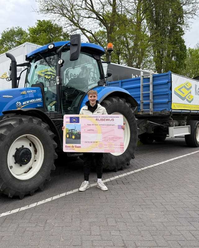 Geslaagd voor het trekkerrijbewijs: cursist in witte trui met rijbewijsbord bij een blauwe lestrekker