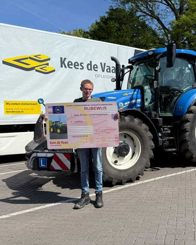 Geslaagd voor het T-rijbewijs: cursist met rijbewijsbord voor een blauwe lestrekker en de lesvrachtwagen van Kees de Vaan