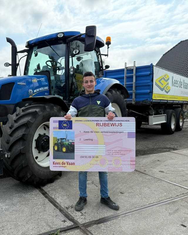 Geslaagd voor het trekkerrijbewijs: cursist in sportjas met rijbewijsbord voor een blauwe lestrekker met kipper